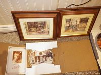 Two framed prints leaning against the wall with three decorative plates on cardboard in front, plus Trish Romance booklet.