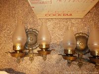 Pair of metal wall sconces with dual frosted glass hurricane-style shades, floral patterned mounting plates, and decorative metal drip trays for candles or lights. Arranged side by side on carpet with vintage cardboard box in background.