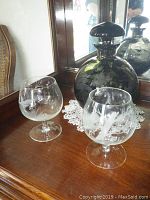 Photo showing the smoky glass decanter with stopper and two clear etched snifters on a wooden surface with lace doily and mirror background