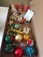Box containing various Christmas ornaments including baubles, figurines, and fabric items arranged in rows.