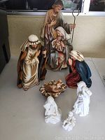 Full set of painted nativity figures arranged on tabletop showing all main characters and two smaller white angel figurines and manger piece.