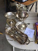 Photo of assorted silver and silver-plated serving pieces on a white table including a lidded pitcher, goblet, standing compote dish, scalloped bowl, serving tray with utensils.