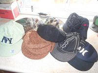 Seven hats laid out on surface: green/cream NY baseball cap, brown newsboy cap, black Tilley newsboy hat, camouflage camp hat, black Boston Red Sox baseball cap, black Hurley logo baseball cap, black winter hat.