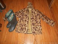 Full view of camouflage Orvis flotation jacket and black Russell waterproof boots with green collar on wooden floor