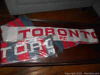 Two knitted Toronto FC scarves displayed on a wooden table, one unpackaged with white and red stripes, the other wrapped in plastic packaging with red, navy blue, and white stripes and Toronto FC text.