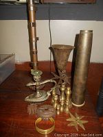 Overview of brass items including bomb casing vase, candlestick holder, chess pieces, bracelet, and other decorative brass pieces on wood floor.