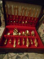 Full open wooden case showing multiple compartments with gold plated cutlery pieces including knives, forks, spoons, and serving utensils arranged on red velvet lining.