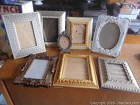 Eight assorted photo frames arranged on a wooden table showing various styles and finishes including silver, bronze, gold, and white.