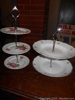 Both vintage tiered dessert stands displayed on a wooden surface against a brick wall background, showing size and floral plate details.