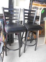 Full view of two black metal bar stools with vinyl seats and ladder backrest.