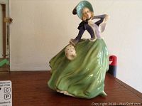 Side profile of Royal Doulton Autumn Breezes figurine showing green skirt, blue jacket, bonnet, and face details.