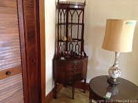 Full view of dark wooden corner cabinet with 4 drawers and open display shelves, standing beside a wooden door and lamp.