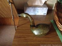 Full side view of greenish-brown signed goose sculpture with raised neck on wooden surface near tissue box.