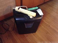 Black Staples paper shredder with attached white surge protector power strip with several outlets, placed on wood floor.