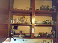 Wooden kitchen cabinet shelves with dishes, glasses, mugs, ceramic tray, and fruit bowls.
