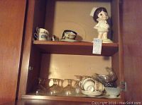Three shelves in wooden cabinet with large ceramic girl chef figurine on top shelf, small cups, clear glass goblets and bowls below