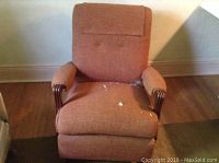 Front view of brown fabric upholstered rocker chair showing seat, cushioned armrests, and wooden arms with visible fabric wear and some damage on the seat.