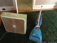Photo of beige Torcan box fan and turquoise Hoover upright vacuum cleaner side by side on carpet.