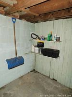 Wide view of basement corner showing blue plastic snow shovel hanging on wall, shelves holding various household chemical containers, toolbox and other items.