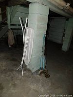 Metal ironing board and Hoover polisher leaning against a gray concrete pillar in a basement area.
