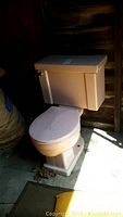 Photo showing side view of pink American Standard toilet with tank and seat, placed on concrete floor near wood wall.