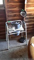 Metal commode with removable bucket, padded seat and armrests, positioned in front of a wooden wall.