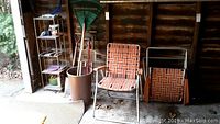 Wide photo showing garden tools standing in a brown cylindrical container next to two vintage lawn chairs with orange and white woven webbing, along with a white shelving unit.