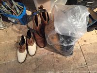 Photo showing men's saddle shoes (two-tone white and brown), brown leather cowboy boots, and a bag containing some of the other items on a wooden floor.