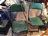 Pair of black folding chairs with green seats and backs, leaning against surfaces with two walking sticks resting between them.