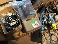 Top view of assorted power cords and computer wires in a cardboard box with a clear plastic zip bag containing additional items, next to a plastic tackle box and antique metal tin.