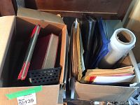 Photo showing box filled with assorted office folders, envelopes, binders, and plastic wrap roll