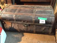 Front and side view of a large vintage wooden chest trunk with metal bands and rivets, showing wear and a detached hinge.