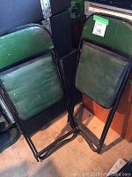 Photo of two green folding chairs folded up and stacked next to each other, showing wear on padding and frames.