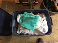 Blue Rubbermaid bin filled with assorted white and green linens, folded and stacked inside.
