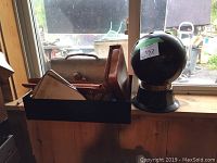 Photo of the black metal bowling ball bar placed on a windowsill with a black box containing assorted vintage items including slide rules, a metal lunch box, pine cones, and shells.