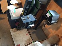 Photo of two pieces of dark room equipment on floor with boxes and chair in background. Shows enlarger bases and lamp heads.