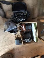 Photo showing several vintage cameras in leather cases, along with camera bags and lenses on a wooden floor.
