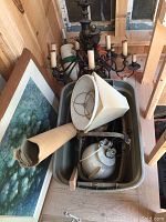 Overview of two metal chandeliers, several lamp shades, a ceramic lamp, antique basket, and framed oil painting leaning against wall