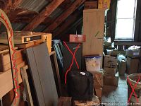 Wide view of attic space showing stacked boxes, wooden panels, and one piece of furniture marked with a red 'X' indicating not for sale.