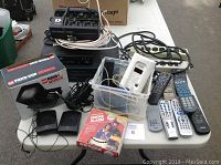 Table with multiple electronics and safety devices including VHS player, DVD player, battery charger, power bars, remotes, speakers, and detectors.