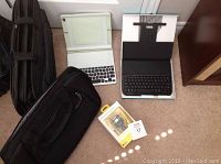Two tablet keyboard folios, two padded computer bags, and digital FM transmitter box arranged on floor.