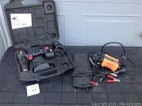 Jobmate cordless drill with battery and charger in carrying case alongside orange solar trickle charger and black MotoMaster 12V battery charger and maintainer with cables.