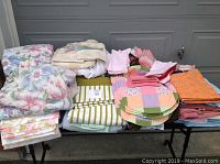 Overview of assorted linens including floral sheet sets, striped sheet sets, placemats, aprons, napkins, and hot pads arranged on tables