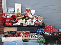 Wide view of table covered with multiple boxes and bags filled with assorted Christmas decorations including ornaments, wrapping paper, plush items, and centerpieces.