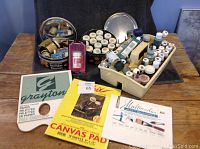 Photo of assorted acrylic paint bottles, glass etching and stain glass supplies in containers, canvas pads and paper palettes on wooden table