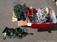 Box with green wire Christmas lights, several portable spotlights with green bases, and a red plastic tote filled with white wire Christmas lights and light timers