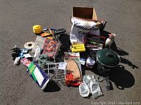 Various camping gear items including stainless steel s'mores grill, grilling rocks, fire logs, pot, and assorted trailer parts and bulbs spread on the ground.