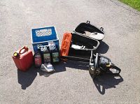 Full lot overview showing the chain saw, carrying case, bar guard, oils, gas can, extra chain, and tools arranged outdoors on asphalt.