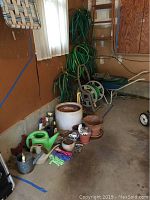 Shows garden hoses coiled on pegboard wall, stacked terra cotta and ceramic pots, watering cans, gloves, small gardening tools on garage floor.