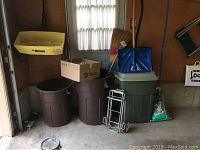 Three brown plastic trash cans: two without lids and one with lid; a yellow Ortho lawn spreader; blue tipped luggage cart; brooms; kitty litter bag on floor in garage corner.